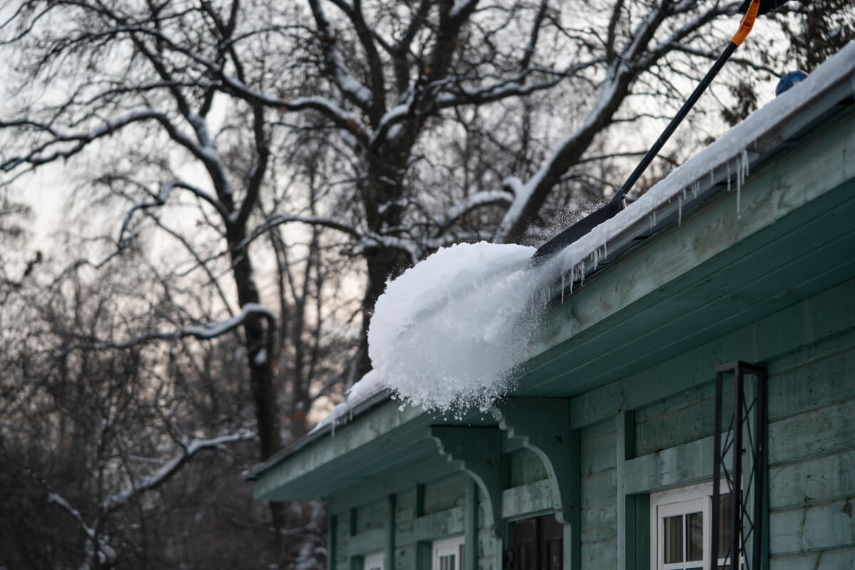 Osoba odśnieża dachy domu, usuwając śnieg łopatą.